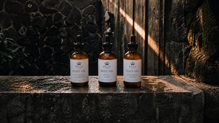 Peace Love & Soap Body Oil collection featuring equal-size Patchouli Lavender, Hotel Luxe, and Baccarat Rouge 4 oz bottles in a moody garden setting