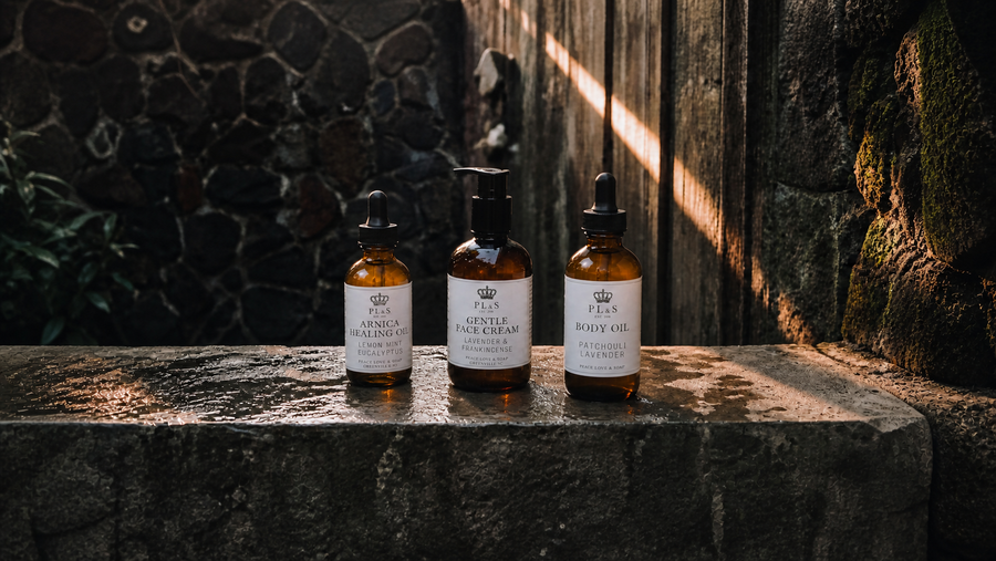 Peace Love & Soap body collection in a moody garden setting, zoomed out with correctly scaled 2 oz Arnica oil and 4 oz body oil